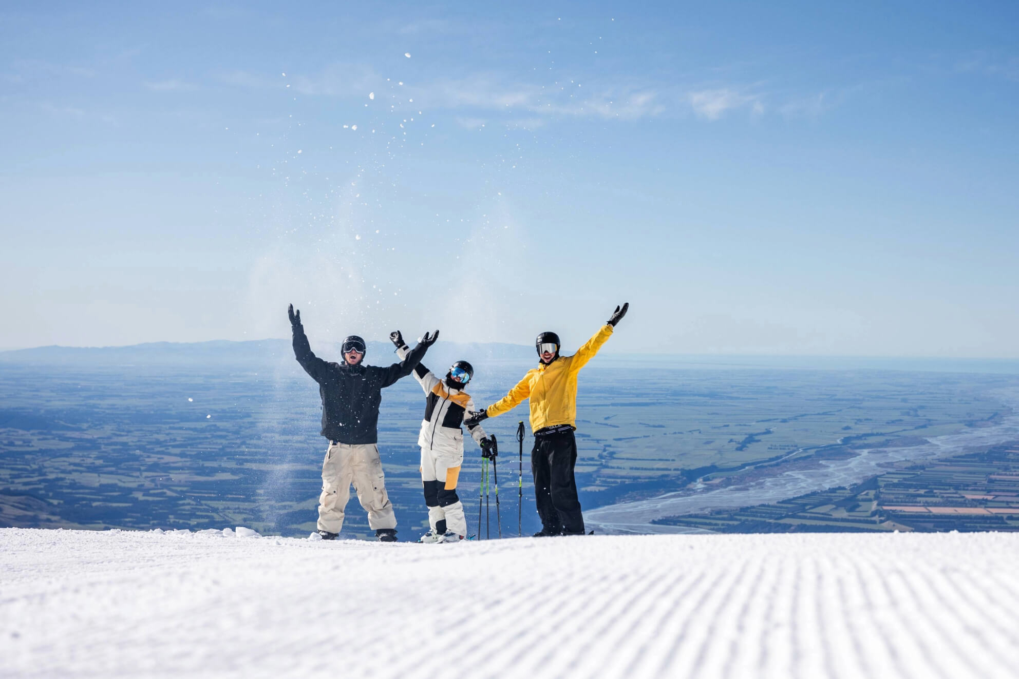 Three skiers in colorful jackets cheering and throwing snow on a sunny mountain slope overlooking a vast valley.