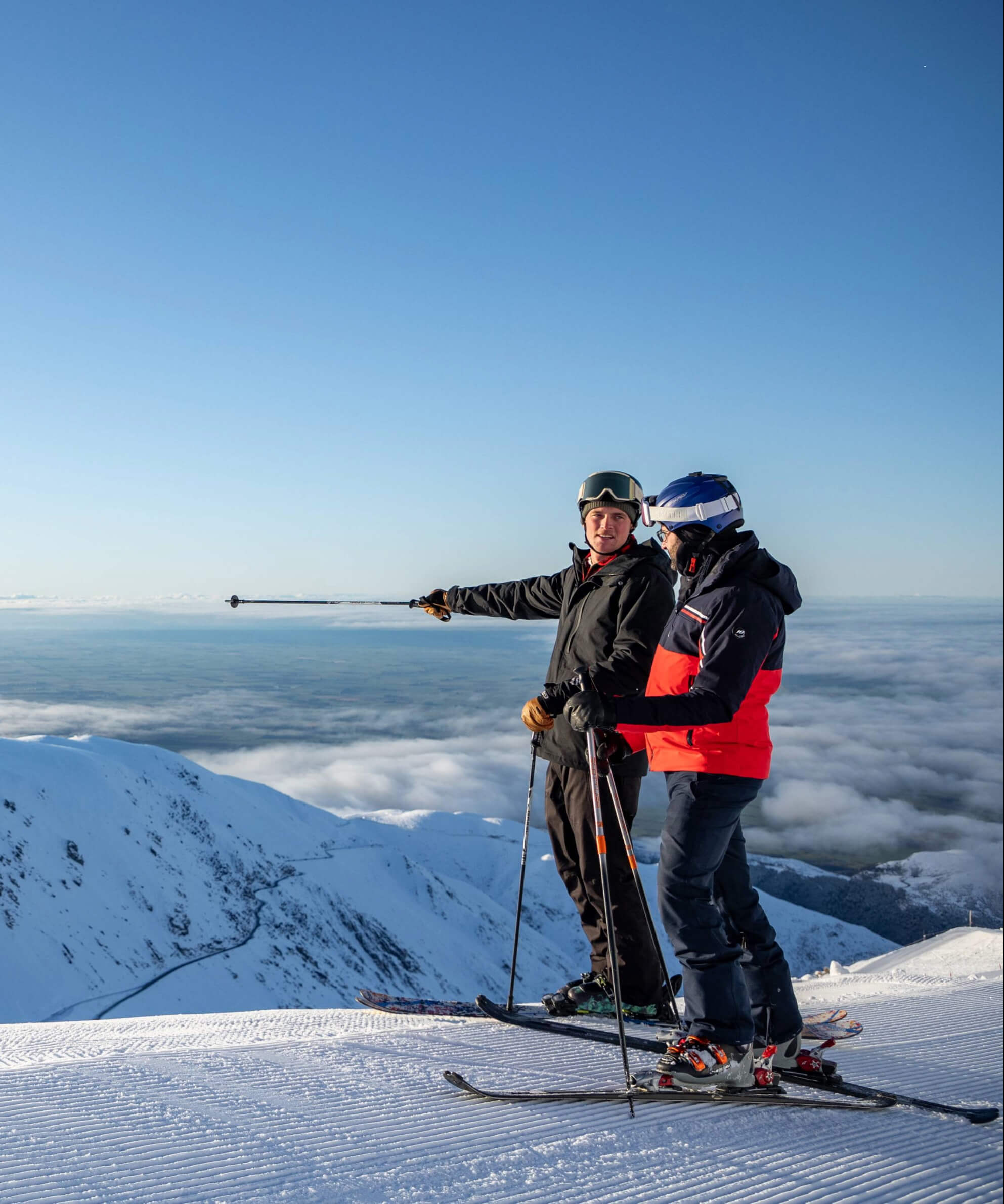 Two skiers standing on a snowy mountain peak with ski poles, one pointing towards the distant view over clouds.