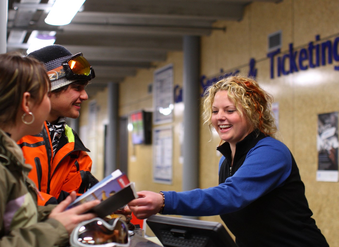Smiling ticket clerk handing a ticket to two customers wearing winter gear at a ski resort counter.
