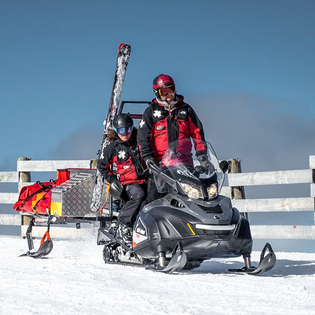 Two ski patrol rescuers on a snowmobile towing rescue supplies on snow.