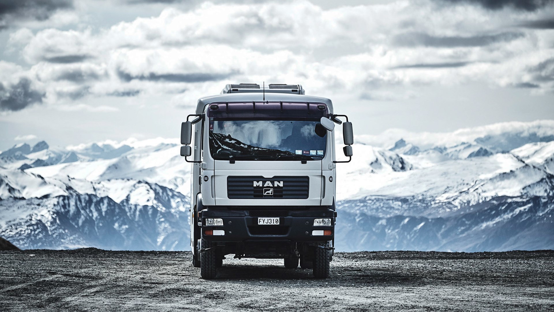 Front view of a parked MAN truck on a gravel surface with snow-covered mountains in the background.