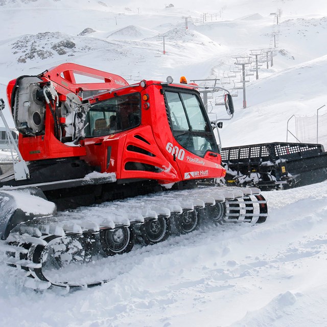 Red snow groomer vehicle on snow-covered mountain with ski lift towers in the background.