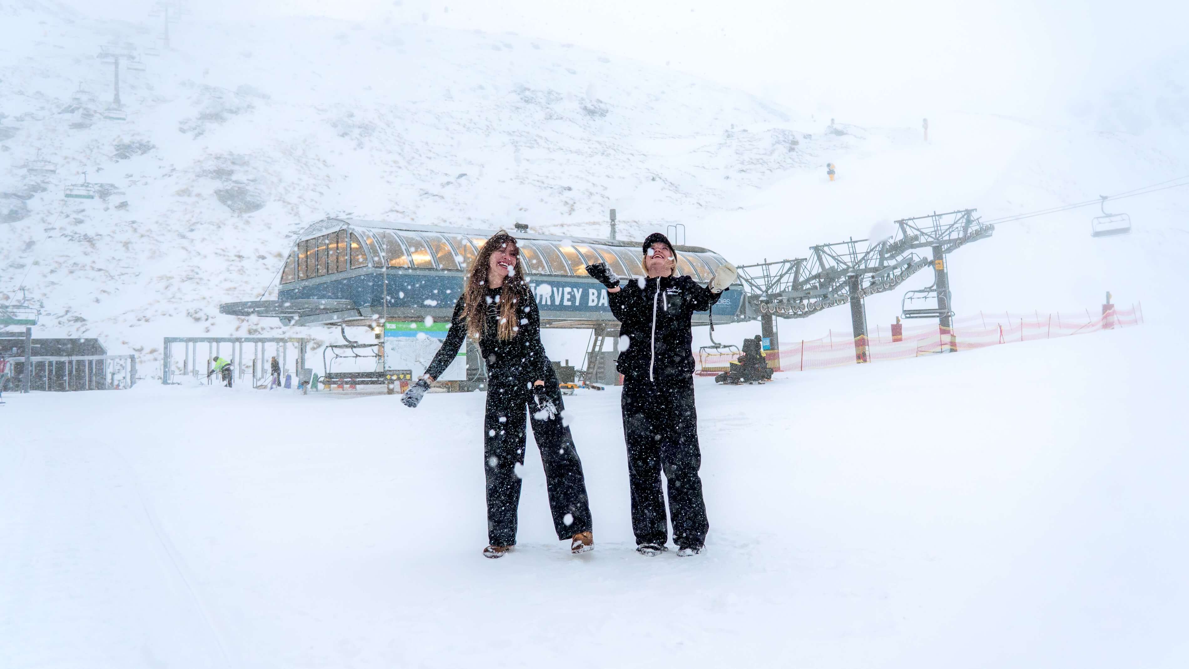 Two people in black winter clothes enjoying heavy snowfall near a ski lift station on a snowy mountain.
