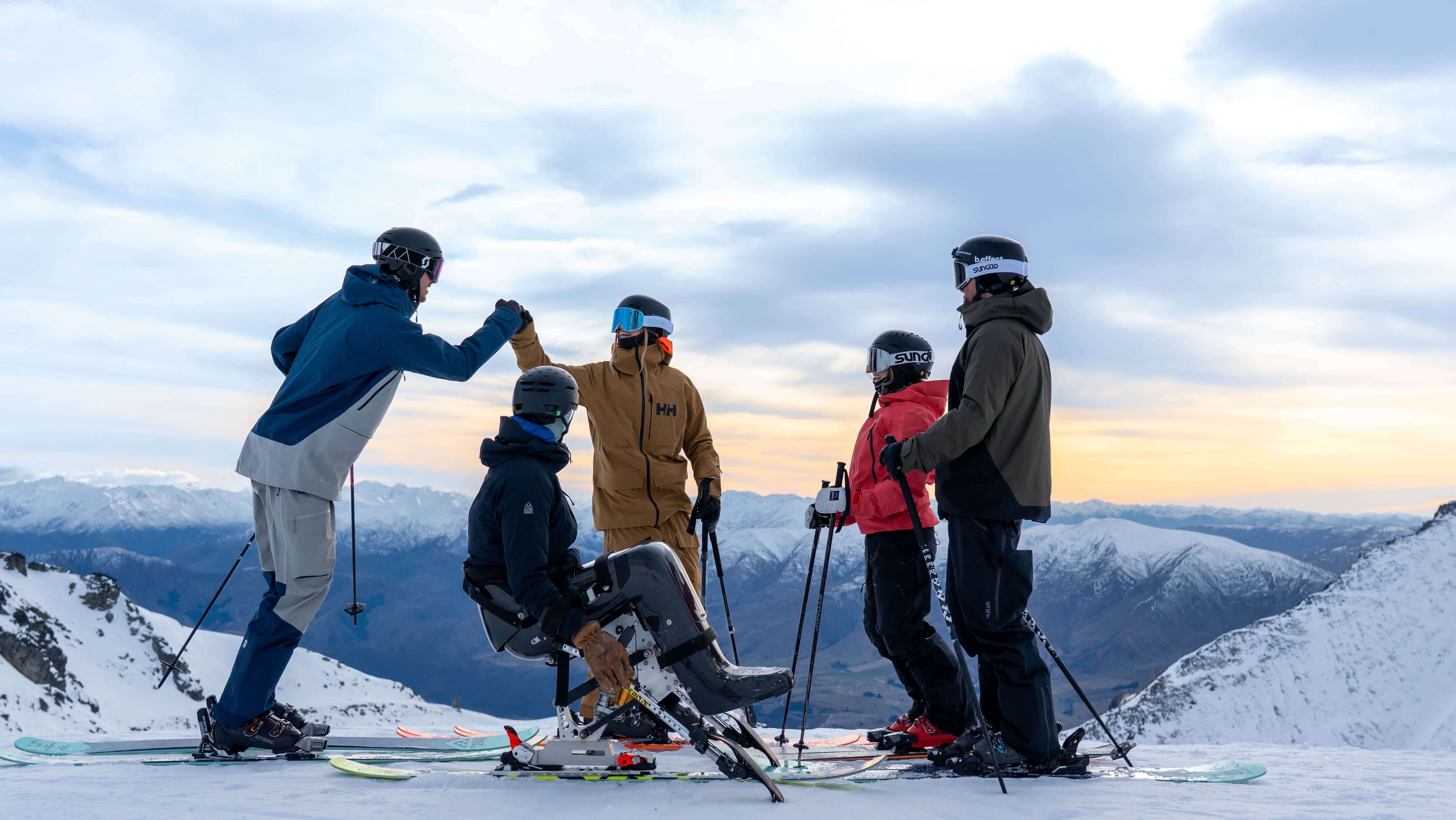 Group of four skiers, including a person with a sit-ski, enjoying a snowy mountain view at sunset.