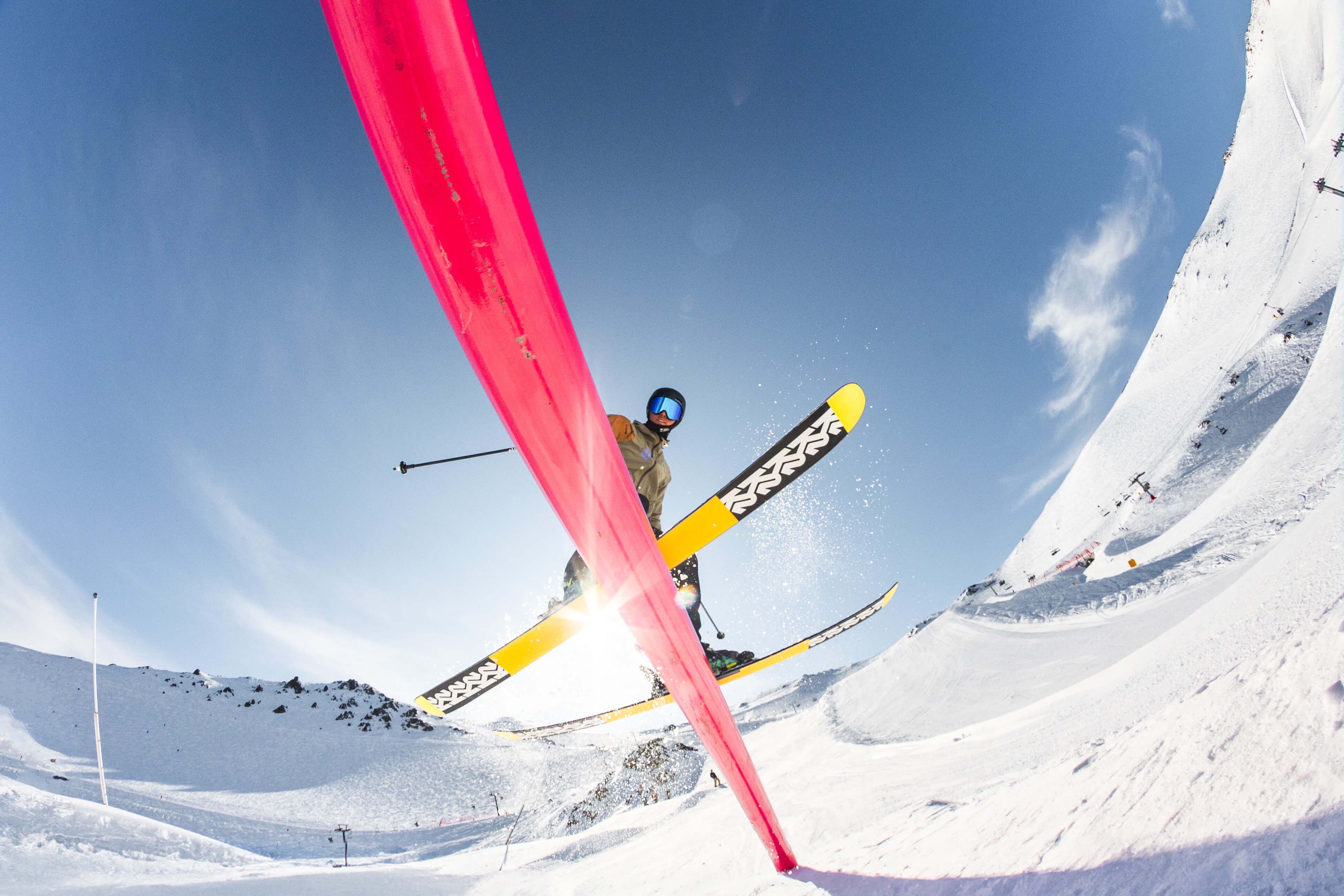 Skier in mid-air jumping over a red rail on a snowy mountain under a clear blue sky.