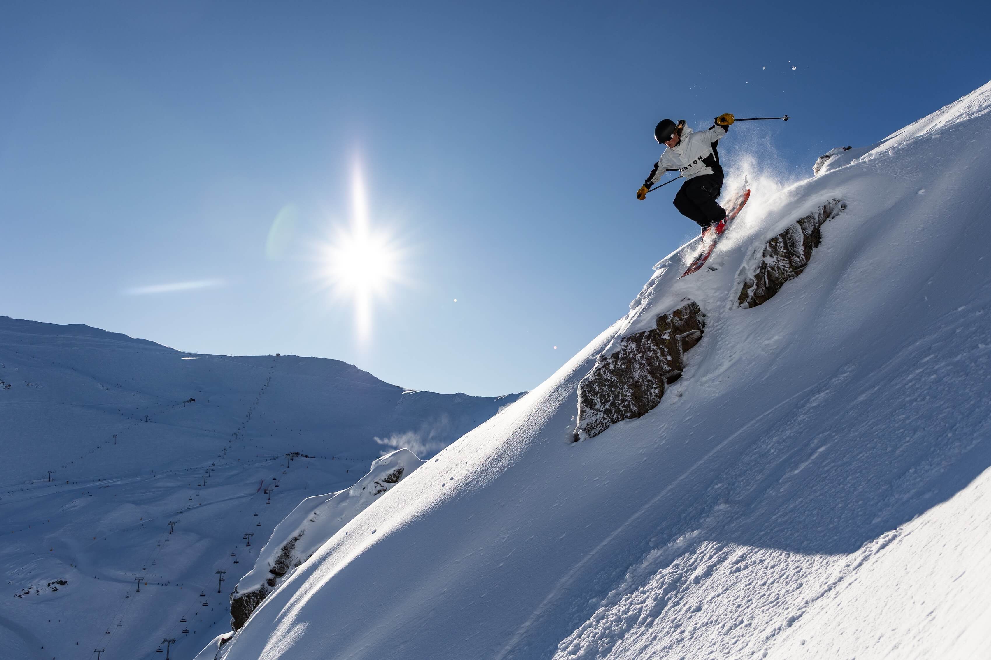 Skier jumping off a snowy cliff on a bright, clear day with mountains in the background.