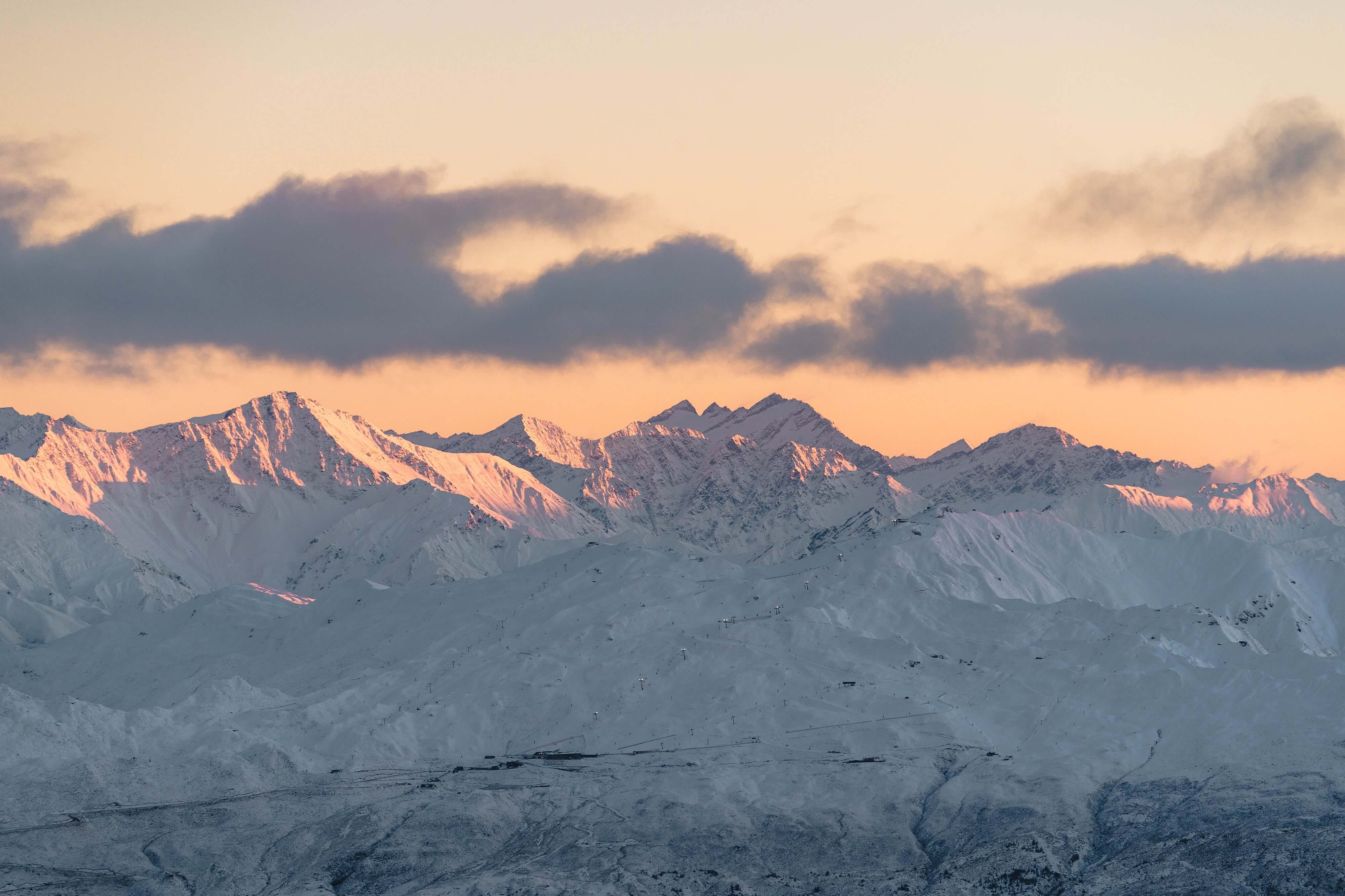 Snow-covered mountain range illuminated by soft orange light under a cloudy sky at sunset.