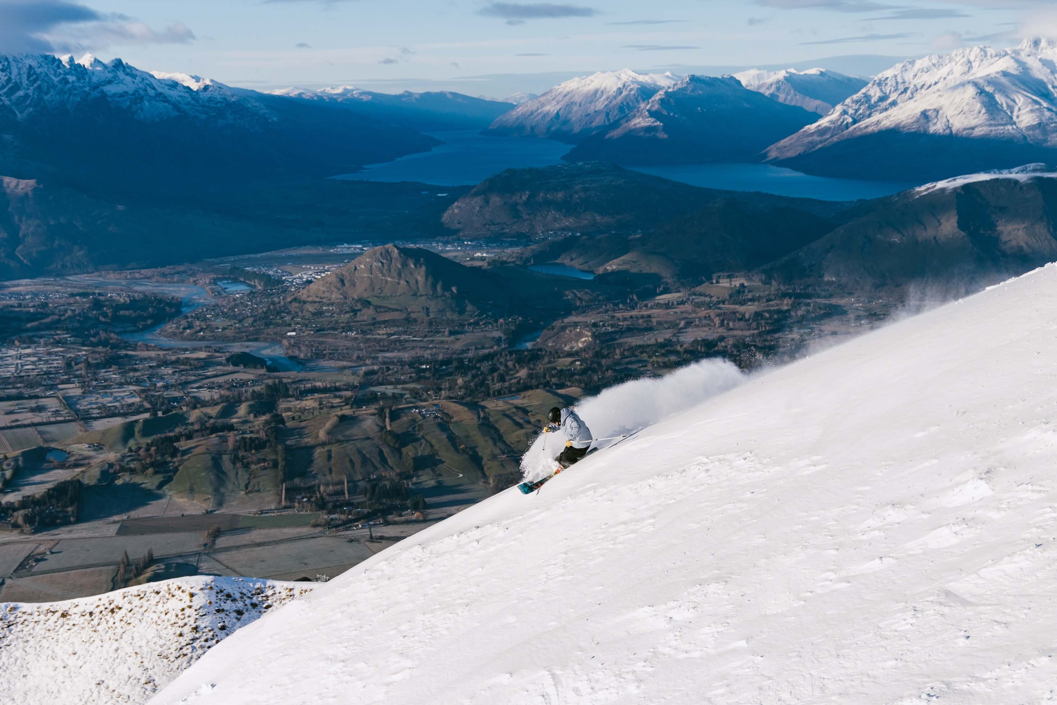 Skier carving down a snowy mountain slope with a valley and snow-capped peaks in the background.