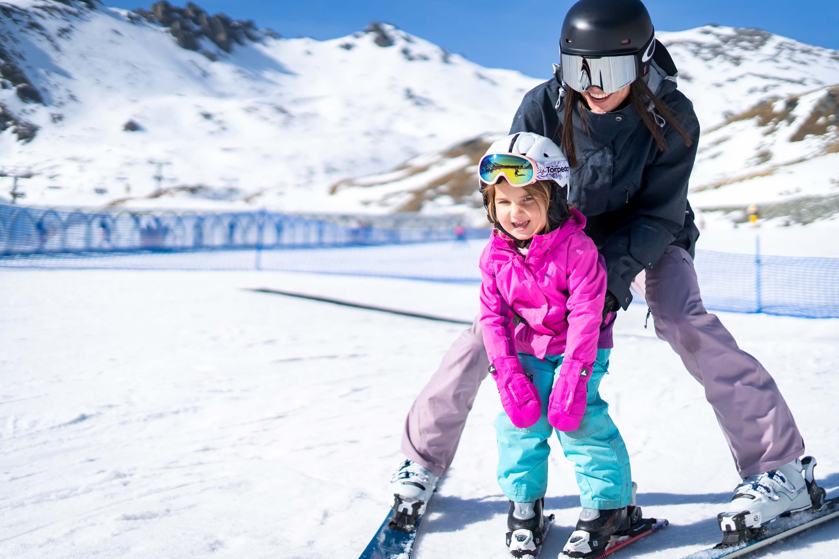 Adult helping child in pink jacket and blue pants ski on snow with mountains in background.