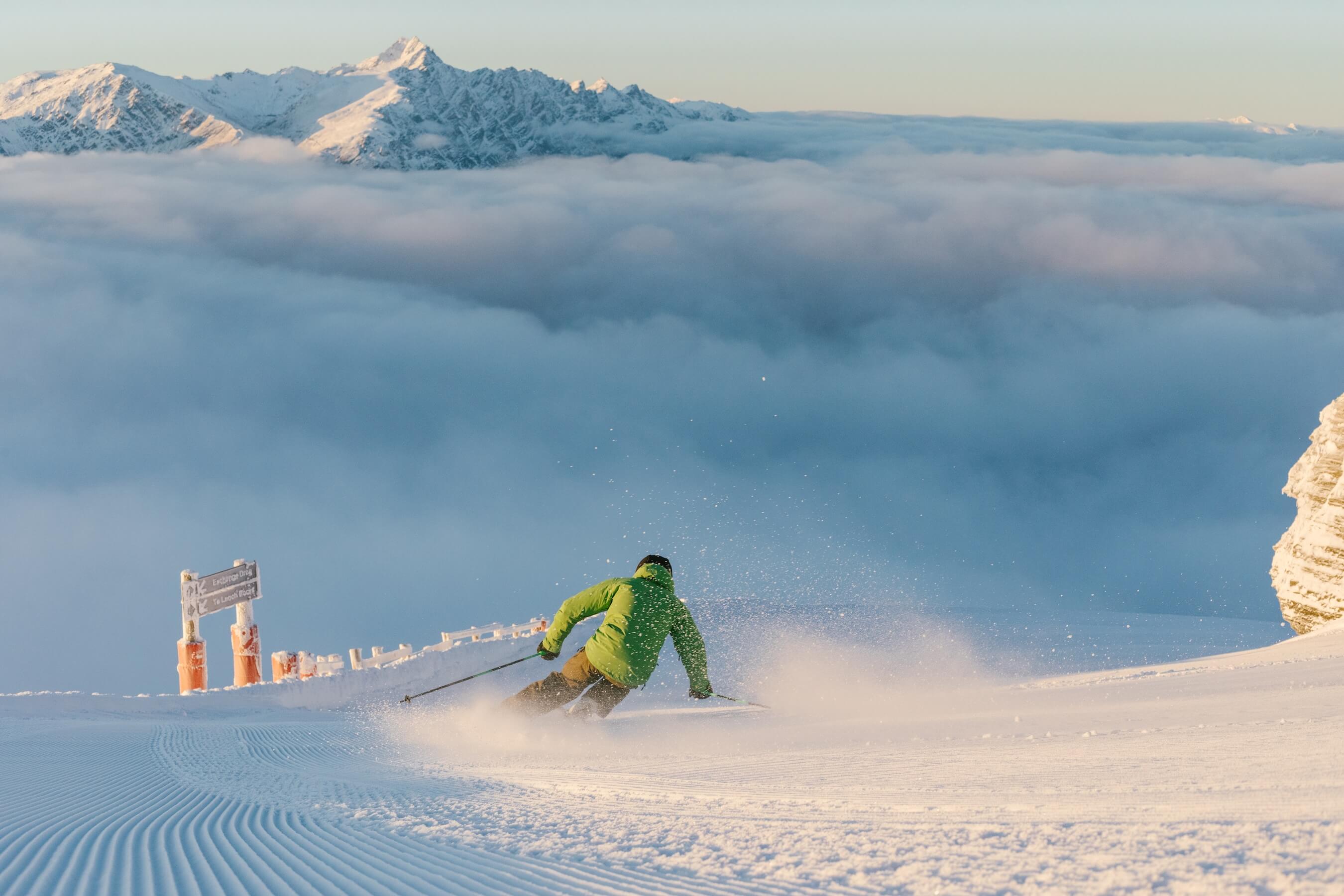 Skier in green jacket carving turns on groomed snowy slope above cloud-covered valley at sunrise.