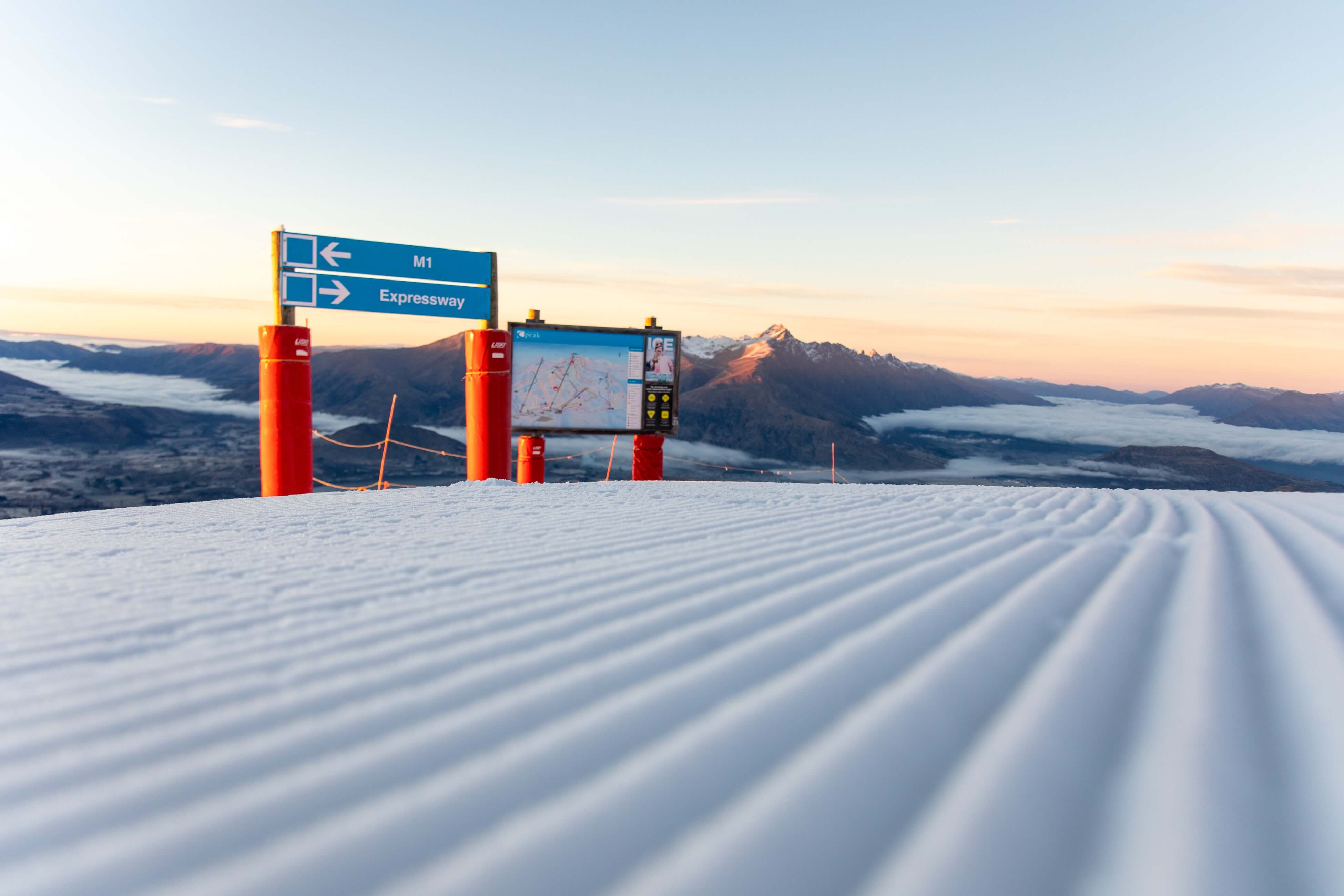 Groomed ski slope with signage and mountain peaks in the background at sunset.