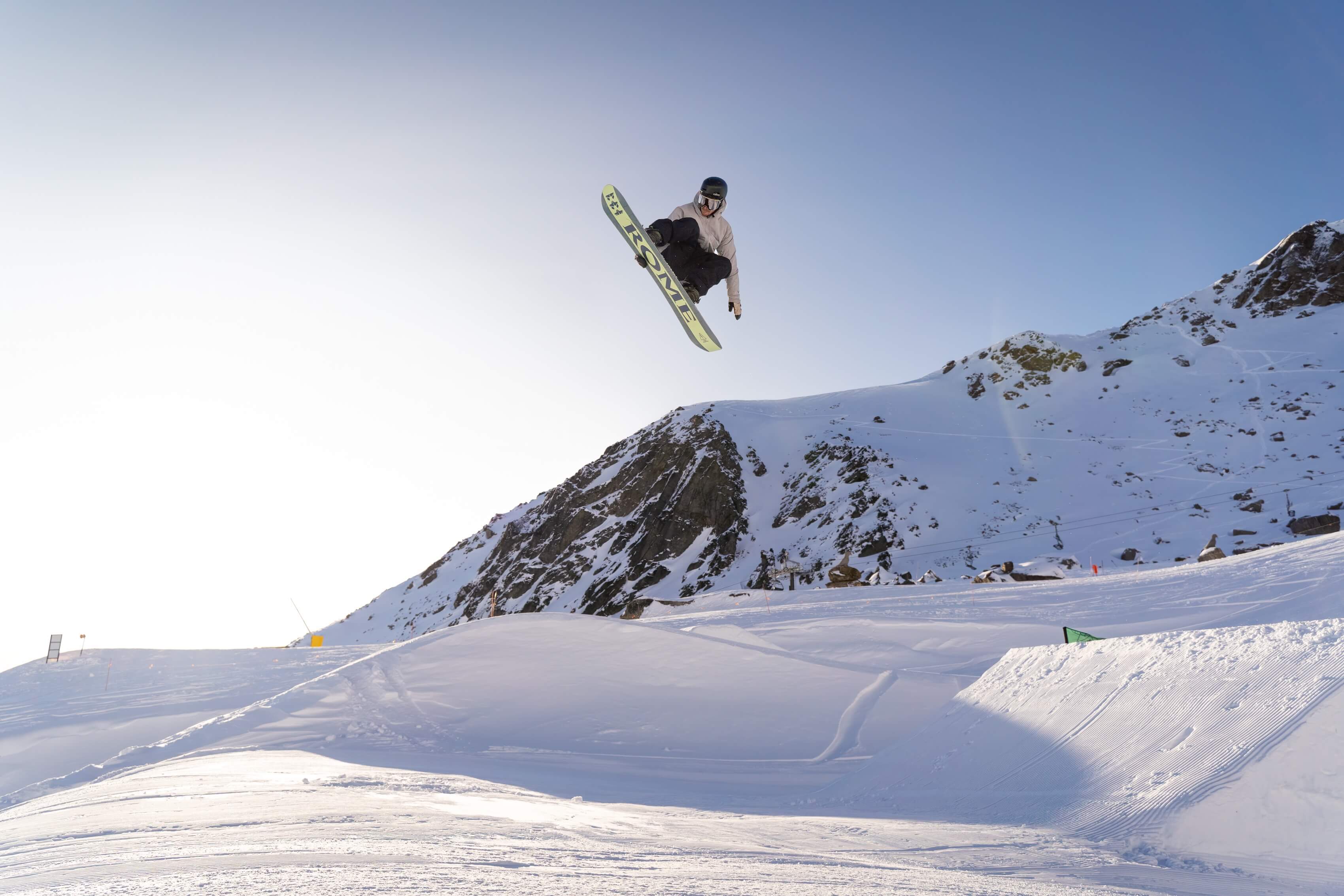 Snowboarder performing a jump mid-air over a snowy mountain slope under clear sky.