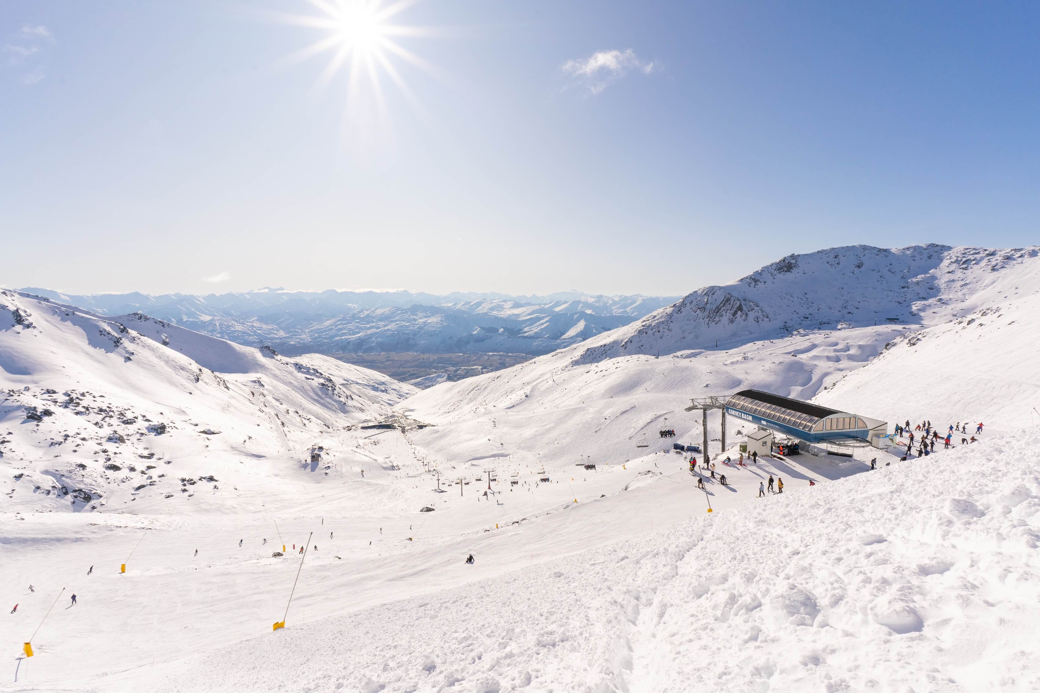 Skiers on snowy mountain slopes near a ski lift station under bright sun and a clear blue sky.